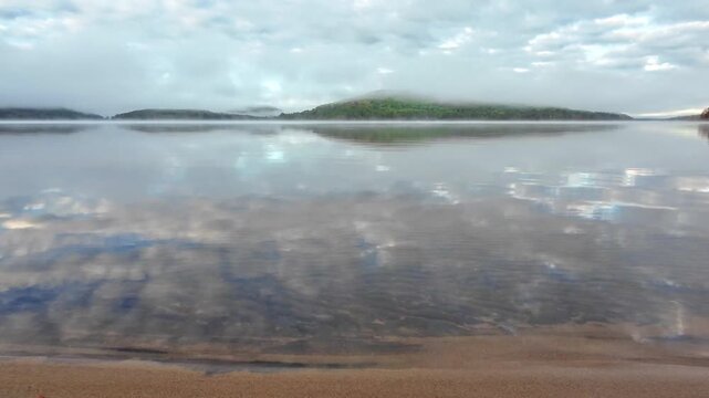 Calm water reflecting clouds above a sandy shoreline on Grand Lake in Achray, Algonquin Provincial Park, Ontario, Canada, with low clouds and fog over the distant forested hills.