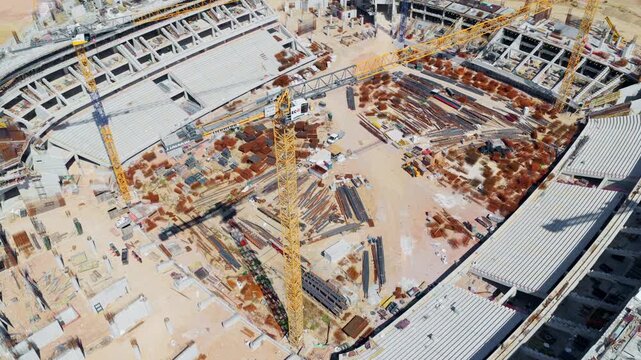 Top-down aerial view revealing the massive construction site of a modern sports stadium, with tower cranes and building materials scattered across the unfinished structure and sandy ground