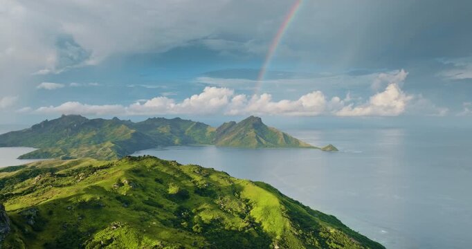 Green mountains and lush tropical landscape of Fiji Islands with a colorful rainbow stretching across the sky, overlooking bays and vast Pacific Ocean under a cloudy sky. Wild nature aerial landscape