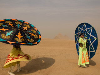 Fototapeta premium Two Egyptian tanoura dancers joyfully spinning vivid folkloric skirts overhead in the desert before the Giza pyramids ideal for cultural tourism and