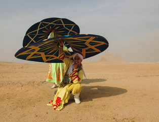 Fototapeta premium Festive Egyptian tanoura dancer spins a colorful patterned skirt overhead and smiles while kneeling in the sandy desert before the ancient Giza
