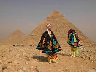 Fototapeta premium Two Egyptian tanoura folk performers in vibrant colorful spinning dance robes standing proudly before the Giza pyramids under bright sun ideal for
