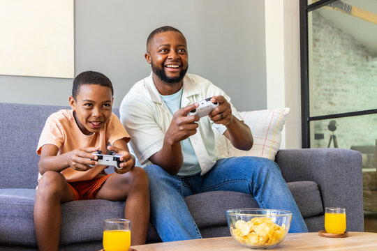 African American dad and preteen son sitting on gray sofa holding white controllers, chips