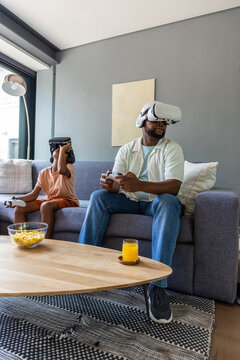 African American father and teenage son holding controllers and wearing headsets on gray sofa