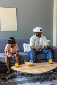 African American dad, teen son wearing VR headsets holding controllers at home with chips, juice