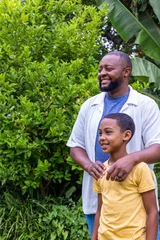 African American father and son standing in backyard garden among banana leaves, hands on shoulders © wavebreak3
