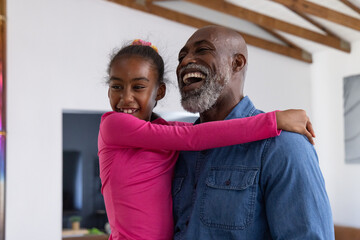 Obraz premium African American senior man and girl laughing, hugging in living room at home with pink top