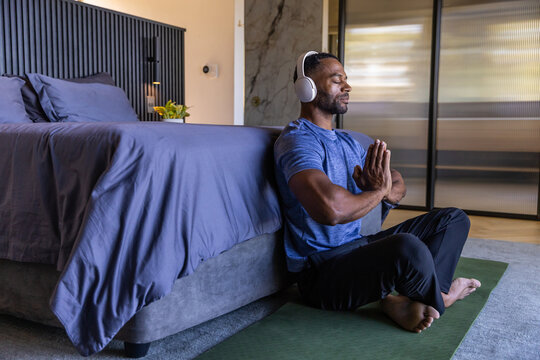 African man meditating on green mat in bedroom, wearing sportswear and headset, copy space