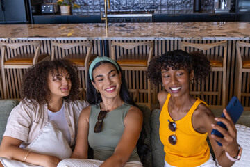 Naklejka premium Diverse female friends sitting on sofa in kitchen holding smartphone smiling wearing sunglasses