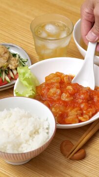 Scooping shrimp with chili sauce (ebi chili) with a spoon on a wooden dinner table, with bang bang chicken (banbanji) in the background, close up, vertical video.