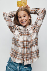 Happy girl smiling with hands behind head holding yellow autumn leaves wearing a plaid shirt and ripped jeans on gray background. Childhood, fall, casual style and joy.