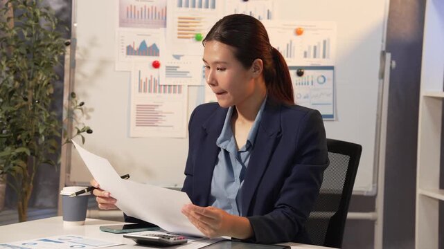 Businesswoman in office reviewing printed reports at desk with charts on wall, coffee cup and calculator nearby, focused and calm during evening work.