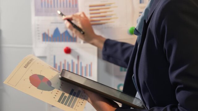 Businesswoman pointing at printed charts on a glass wall during a meeting, showing sales data, planning, analysis and office teamwork in a modern workplace.