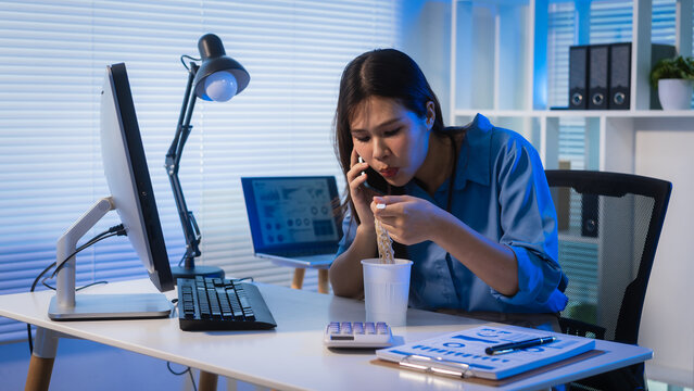 Young businesswoman eating instant noodles at her desk while talking on the phone in a modern office at night, showing overtime work, busy schedule, and office stress.