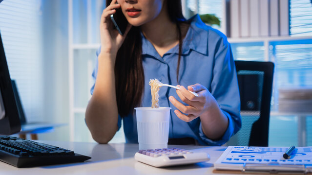 Young businesswoman eating instant noodles at her desk while talking on the phone in a modern office at night, showing overtime work, busy schedule, and office stress.