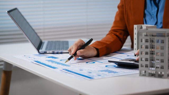 Business professional reviewing financial charts at a desk while holding a small building model, suggesting real estate investment, planning, and market analysis.