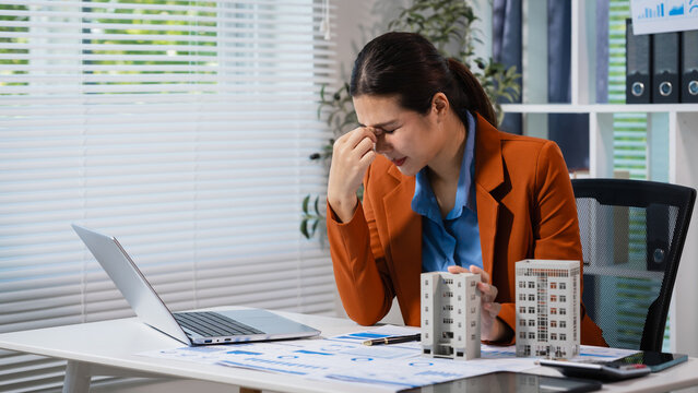 Stressed businesswoman at office desk looking at laptop and paperwork, holding her nose in frustration, with small building models and calculator nearby.