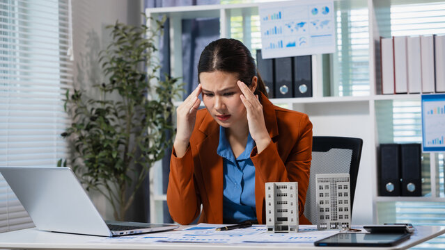 Stressed businesswoman at office desk looking at laptop and paperwork, holding her nose in frustration, with small building models and calculator nearby.