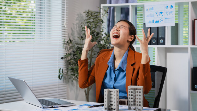Excited businesswoman at office desk celebrating success with laptop, charts and small building models, showing joy, achievement and positive energy in a modern workplace.