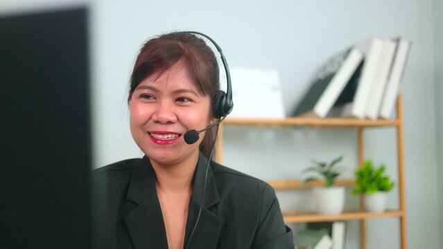 Professional Asian female customer service operator wearing a headset working on a computer, talking happily with clients in a friendly remote workspace office environment.