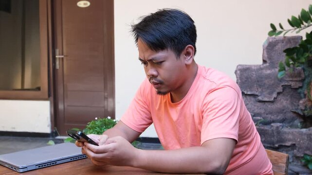 A frustrated Asian man typing on smartphone while arguing in chat or social media beside a laptop outdoors.