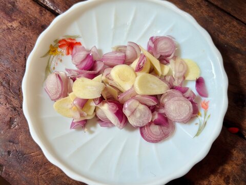 Top view of sliced raw shallots and garlic cloves served on a white floral ceramic plate over a rustic wooden table background, capturing fresh cooking ingredients and aromatic spices.