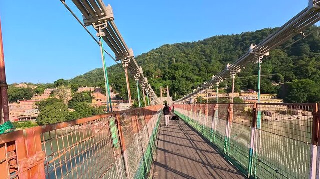 Walking perspective down the center of a pedestrian steel suspension Ramjhula bridge over the Ganges River