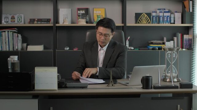 A businessman in a suit sitting at a modern executive desk reviewing documents under warm lamp light with bookshelves in the background at night.