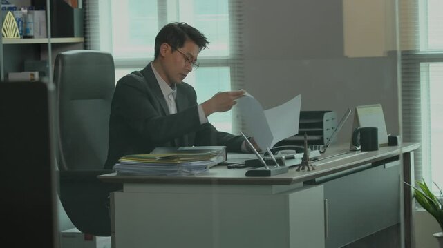 A businessman in a suit sitting at a modern office desk reviewing documents and files stacked on the desk in a bright corporate office environment.