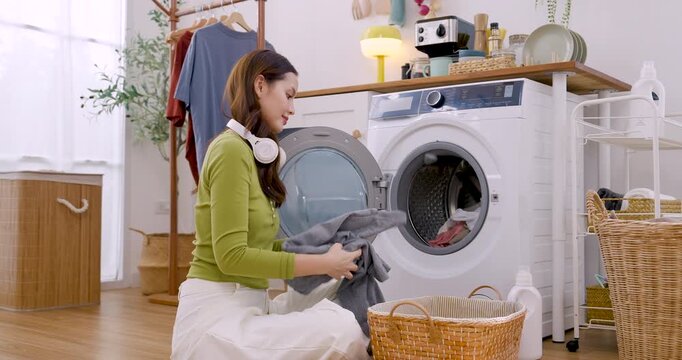 Young Asian woman sits beside laundry basket, gathers white textile, reaches toward washer, prepares fresh load, creating clean home routine in bright apartment with cozy domestic mood today