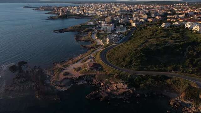 Curving coastal road leads toward the seaside town of Alghero as a drone glides above rocky shoreline at golden hour.