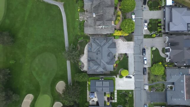 Birds-eye aerial perspective slowly pans sideways over residential culdesac street showing luxury homes manicured yards swimming pool adjacent green golf course sand bunkers under daylight