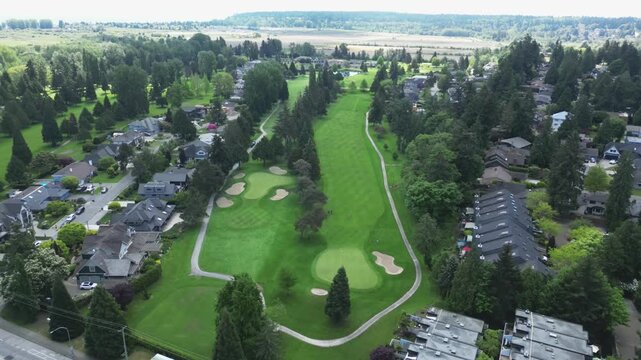 High-angle aerial perspective moves forward showing an extensive green golf fairway with multiple sand bunkers flanked by residential houses and modern townhomes stretching toward distant farmlands