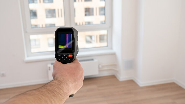 Hand holds thermal imaging device. Screen shows heat patterns in color. Room has light wood floors and white walls. Window reveals city buildings outside. Check for energy leaks or insulation problems