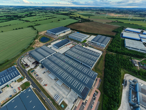 Aerial view of a modern logistics park with large warehouse buildings, loading docks, and parking areas surrounded by green fields in Biggleswade, England, United Kingdom.