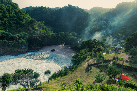 High-angle stock photo capturing a red camping tent, a small coastal mosque, and smoke rising over the lush bay of Ngitun Beach.