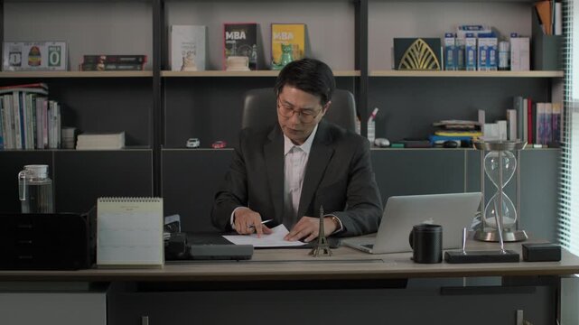 A businessman in a suit sitting at a modern executive desk writing notes beside a laptop with bookshelves and decorative items in the background.