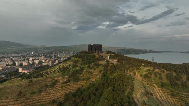 Drone shot capturing the historical Chronicle of Georgia monument complex during a scenic evening sunset. The huge stone pillars are set on a green hill next to the calm Tbilisi Sea reservoir