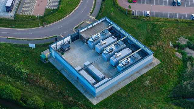 Aerial view of an industrial utility site with machinery units on a concrete pad, surrounded by green grass and a nearby parking lot in Biggleswade, England, United Kingdom.