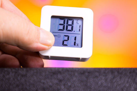 Close-up shot of a digital thermometer showing an extreme temperature of 38.1C with a very low humidity level of 21%. A hand holds the compact device against a vibrant, warm-colored blurred background