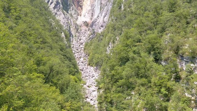 Boka Waterfall cascades down a massive rocky cliff in the scenic Soca Valley, Slovenia, surrounded by lush green forests and a wide boulder stone canyon during a bright, sunny beautiful summer day.