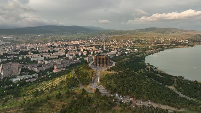 Drone shot capturing the historical Chronicle of Georgia monument complex during a scenic evening sunset. The huge stone pillars are set on a green hill next to the calm Tbilisi Sea reservoir