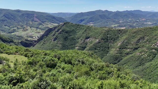 Ampio paesaggio montano ricoperto di vegetazione verde, con valli e creste che si estendono in lontananza sotto un cielo azzurro limpido. Scena naturale vasta e luminosa.