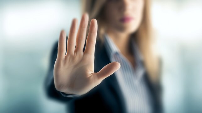 Professional business woman wearing a dark blazer extends her hand forward in a clear gesture to stop or block unwanted actions while working inside an office, showing rejection