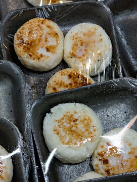 Round cheesecakes with a lightly browned top are individually wrapped in plastic and placed in gray trays. The cheesecakes appear freshly baked and ready for sale in a market or store setting. The pac
