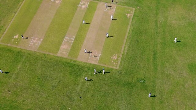 Men playing cricket at Chadwell Heath park, London, England, seen from high drone orbit showing pitch strips, field positions, and summer grass in suburban open space.