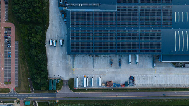 Aerial view of a large industrial warehouse with solar panels on the roof, loading docks with parked trucks, and an adjacent parking lot in Biggleswade, England, United Kingdom.