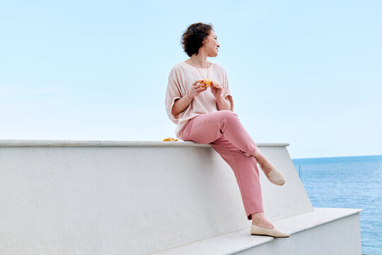 Woman joyfully eating a mandarin by the pool in a pink casual outfit