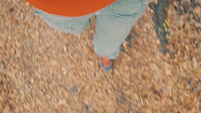 A woman walks through an autumn forest, her feet in blue sneakers crunching on fallen leaves. The ground is covered in a layer of brown and gold foliage, highlighting the changing season.