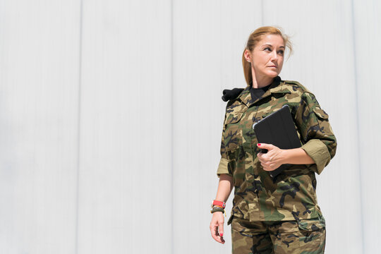 Mature blond female army soldier holding digital tablet while looking away at military base on sunny day